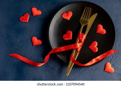 Valentines Day Table Setting On A Dark Blue Background. Black Plate, Golden Cutlery And Red Hearts, Romantic Dinner Concept