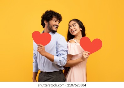 Valentine's holiday. Portrait of indian lovers holding red paper hearts and looking at each other, standing on yellow studio background. Romantic relationship concept