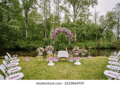Valmiera, Latvai - May 23, 2025 - Romantic outdoor wedding setup with a floral arch, white chairs, and flower arrangements by a calm forest riverbank.