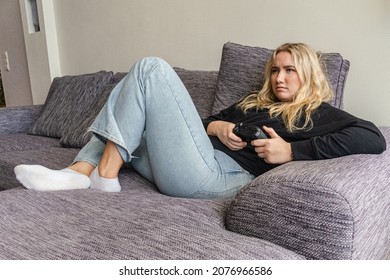 Valmiera, Latvia, 6 November, 2021: Women With Baggy Jeans Sitting In Couch And Playing Video Games. Blonde Girl Wearing Blue Jeans, Black Sweater. Enjoying Games On A Sunny Day.