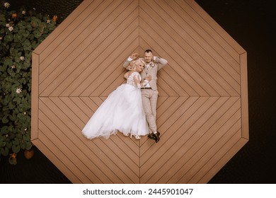 Valmiera, Latvia - Augist 13, 2023 -A bird's-eye view of a bride in a white dress and groom in a beige suit laying down on an octagonal wooden pier, with dark water and lily pads nearby. - Powered by Shutterstock