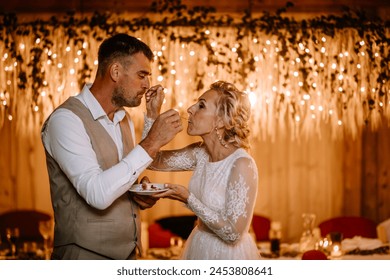 Valmiera, Latvia - August 10, 2023 - Bride and groom feeding each other cake with twinkling lights in the background. - Powered by Shutterstock