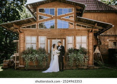 Valmiera, Latvia - August 15, 2024 -  A bride and groom stand together outside a rustic wooden house, gazing at each other on their wedding day. - Powered by Shutterstock