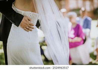 Valmiera, Latvia - August 15, 2024 - Close-up of a groom holding the bride’s waist during their wedding, focusing on their embrace and wedding attire with guests blurred in the background. - Powered by Shutterstock