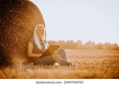 Valmiera, Latvia - August 17, 2024 - 
A woman sits leaning against a hay bale in a field, holding a tablet and looking away thoughtfully in the warm glow of the sunset. - Powered by Shutterstock