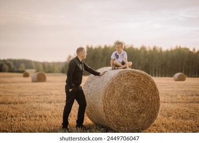 Valmiera, Latvia - August 17, 2024 - A man in business attire smiles at a young boy sitting atop a hay bale in a field at dusk. - Powered by Shutterstock