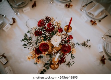 Valmiera, Latvia - August 19, 2023 - Top-down view of a wedding table centerpiece featuring red and white roses, baby's breath, and other flowers, with a golden candle holder. - Powered by Shutterstock