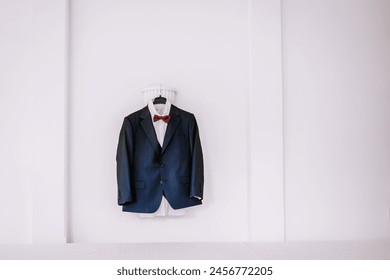 Valmiera, Latvia - August 19, 2023 - A navy blue suit with a white shirt and red bow tie, hanging on a white hanger against a clean white background. - Powered by Shutterstock