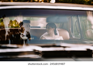 Valmiera, Latvia - August 19, 2023 - A bride and groom sit side by side in a classic car, exchanging a glance with a blurred background. - Powered by Shutterstock