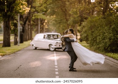 Valmiera, Latvia - August 19, 2023 - Groom lifting bride in his arms on a road, both smiling, with a vintage car and trees in the background. - Powered by Shutterstock