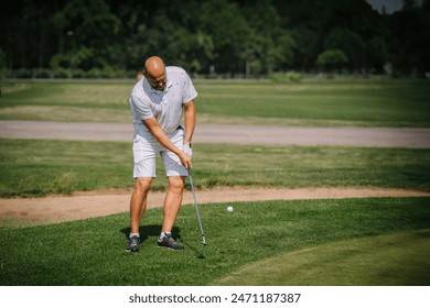 Valmiera, Latvia - June 3, 2024 - A man in a white polo shirt and shorts swings a golf club on a golf course, focusing on his shot on a sunny day. - Powered by Shutterstock