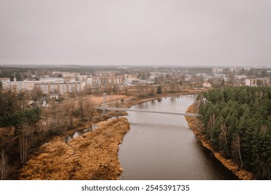 Valmiera, Latvia - November 14, 2024 - Aerial view of a suspension bridge over a river with an urban neighborhood in the background, surrounded by autumn trees under an overcast sky. - Powered by Shutterstock