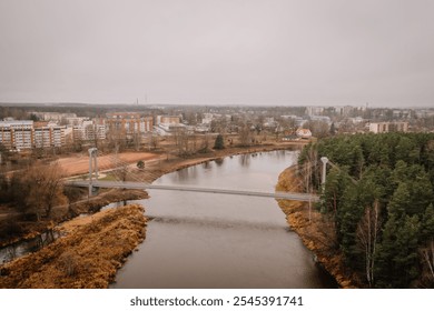 Valmiera, Latvia - November 14, 2024 - Aerial view of a suspension bridge over a river with an urban neighborhood in the background, surrounded by autumn trees under an overcast sky. - Powered by Shutterstock