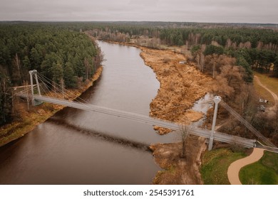 Valmiera, Latvia - November 14, 2024 - Aerial view of a suspension bridge crossing a calm river surrounded by a forest and autumnal landscape, creating a serene and natural scene. - Powered by Shutterstock