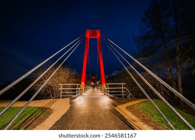 Valmiera, Latvia - November 15, 2024 -  Symmetrical view of an illuminated pedestrian bridge with vibrant red lighting, set against a deep night sky, with cables converging toward the structure. - Powered by Shutterstock