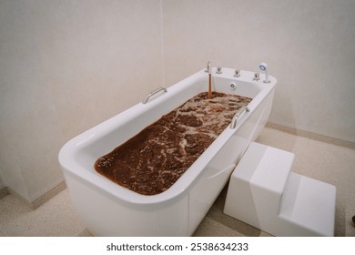 Valmiera, Latvia - October 30, 2024 - A white bathtub filling with bubbling, brown therapeutic water, featuring handrails and a step stool, in a clean, minimalist spa or wellness setting. - Powered by Shutterstock