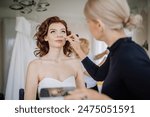 Valmiera, Latvia - September 9, 2023 - Bride getting her makeup done by a makeup artist, preparing for the wedding, with her wedding gown hanging in the background.