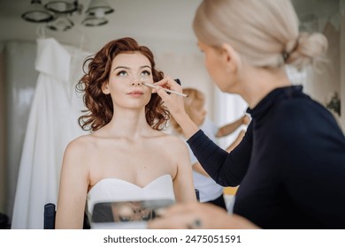 Valmiera, Latvia - September 9, 2023 - Bride getting her makeup done by a makeup artist, preparing for the wedding, with her wedding gown hanging in the background. - Powered by Shutterstock