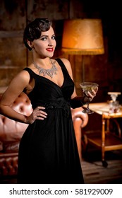 Vamp lady in black dress posing with her hand on hip. Beautiful woman with red lips looking at camera and holding glass of alcohol cocktail.