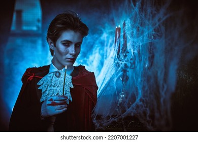 Vampires. Portrait Of A Handsome Young Vampire Standing In His Abandoned Castle In An Elegant 19th-century Clothes And Red Velvet Cloak. Halloween. 