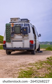 Van loaded with boxes, jerry cans, shovel, and traction boards rests in peaceful grassy field. Scene evokes a sense of adventure and exploration in nature.
