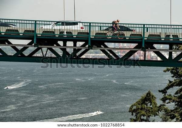 Vancouver, British Columbia,\
Canada - June 30, 2016:  Side view of traffic and cyclist on Lions\
Gate Bridge deck over Burrard Inlet with Vancouver Harbour in\
background.