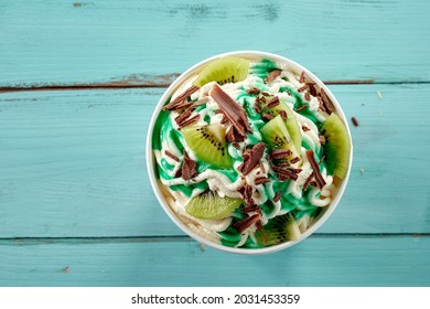 Vanilla Spaghetti Ice Cream Sundae With Fresh Kiwi Fruit And Chocolate Flakes Drizzled With Green Syrup Viewed Top Down In A Tub Over Blue Wood With Copyspace For Menu Advertising