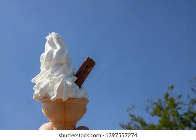 Vanilla white cold Ice cream and ice cream cone with a chocolate flake sticking out the side taken on a sunny day showing the clear blue sky and a few trees blurred in the background. - Powered by Shutterstock