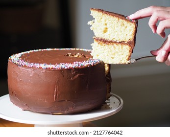 Vanilla Yellow Birthday Cake With Chocolate Buttercream Frosting And Rainbow Sprinkles. Hand And Spatula Lifting Slice From Rest Of Cake.