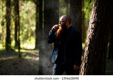 Vape Bearded Man. An Adult White Short-haired Man With A Very Long Beard In Glasses Smokes An Electronic Cigarette Outside In The Dark Forest. Bad Habit That Is Harmful To Health.