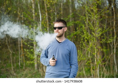 Vape man. An adult white bearded man in glasses smokes an electronic cigarette outside in the forest in sunny day. Bad habit that is harmful to health. - Powered by Shutterstock