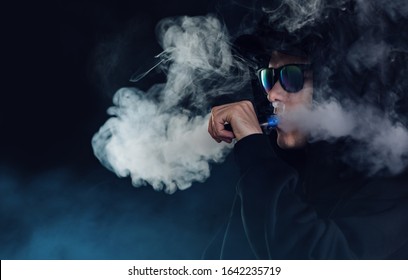vape man. portrait of a handsome guy in a black cap and glasses vaping and exhaling a cloud of vapor from an e cigarette dark background - Powered by Shutterstock