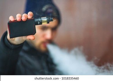 Vape Man. Portrait Of A Handsome Young White Guy In Black Waistcoat And Modern Cap Vaping An Electronic Cigarette And Letting Out Puffs Of Steam