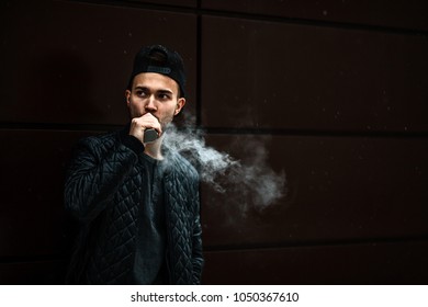 Vape Man. Portrait Of A Handsome Young White Guy In A Modern Black Cap Vaping And Letting Off Puffs Of Steam From An Electronic Cigarette Opposite The Futuristic Urban Background.