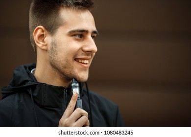 Vape Teenager. Handsome Young White Guy In Black Jacket Vaping An Electronic Cigarette Opposite The Futuristic Urban Background In The Spring. Lifestyle.