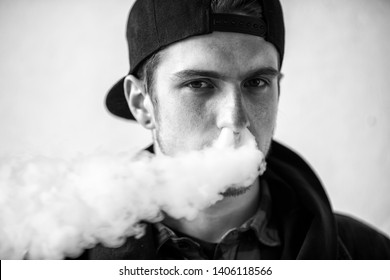 Vape teenager. Portrait of young handsome guy in a cap smoking an electronic cigarette on the street in the spring. Bad habit that is harmful to health. Vaping activity. Black and white. Close up.