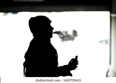 Vape Teenager. Silhouette Of A Young Girl In A Hat Smoking An Electronic Cigarette. Harmful Habit That Is Harmful To Health. Vaping Activity.