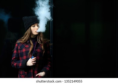 Vape Teenager. Young Pretty White Girl In Checkered Coat And Black Cap Smoking An Electronic Cigarette Opposite Glass Building On The Street In The Spring. Bad Habit. Vaping Activity.