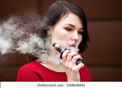 Vape teenager. Young pretty white caucasian girl in a red sweater smoking an electronic cigarette opposite modern brown background on the street in the winter. Deadly bad habit.