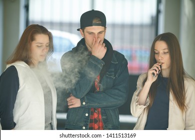 Vape Teenagers. Two Young Cute Girls In Casual Clothes Smoke Electronic Cigarettes Near Nonsmoking Guy Outdoors In The Street In Summer Day. Bad Habit That Is Harmful To Health. Vaping Activity.