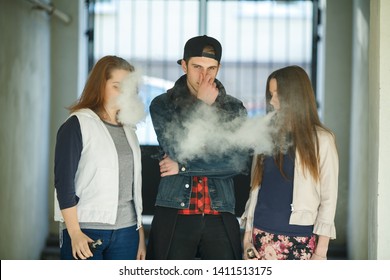 Vape Teenagers. Two Young Cute Girls In Casual Clothes Smoke Electronic Cigarettes Near Nonsmoking Guy Outdoors In The Street In Summer Day. Bad Habit That Is Harmful To Health. Vaping Activity.