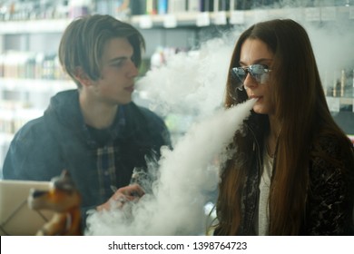 Vape Teenagers. Young Cute Girl In Sunglasses And Young Handsome Guy Smoke An Electronic Cigarettes In The Vape Bar. Bad Habit That Is Harmful To Health. Vaping Activity.