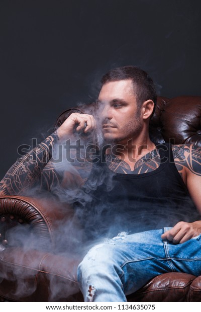 Vaper. The man with\
tattoos sits on a leather sofa smoke an electronic cigarette on the\
dark background.