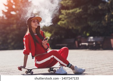 Vaping girl. Young woman sitting on skateboard and vape e-cig. Pretty young female in black hat, red clothing vape ecig, vaping device at the sunset. Toned image. Hip-hop style.