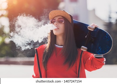 Vaping girl. Young woman with skateboard vape e-cig. Pretty young female in black hat, red clothing vape ecig, vaping device at the sunset. Toned image. Hip-hop style.