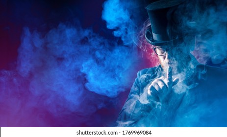 Vaping. Vaper In A Top Hat In A Cloud Of Smoke. A Man Smokes An Electronic Cigarette On A Dark Background. A Man In A Top Hat Puffs Out Smoke From An E-cigarette. Place For Text.