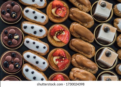 Variety Of Delicious Confectionery Products On Black Tray. A Mix Of Chocolate Mousse, Eclair Cakes And Fruit Tarts For A Candy Bar, Or A Pastry Shop Showcase Presentation. Flat Lay, Top View Dessert.