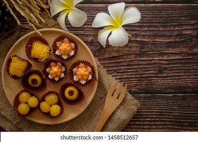 a variety of elegantly beautiful Thai Desserts (Khanom Wan Thai) serving in a wooden plate on a dark wooden table. The bites in gold color and different flower shapes.Thai dessert, Auspicious dessert 