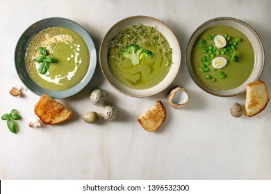Variety of green vegetable soup asparagus, broccoli and pea, decorated by greens, vegetables, quail eggs, cream, olive oil, in ceramic bowls in row over white marble background. Flat lay, space