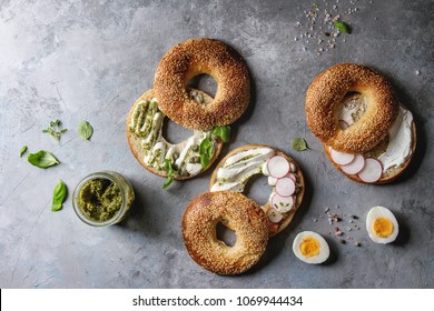 Variety Of Homemade Bagels With Sesame Seeds, Cream Cheese, Pesto Sauce, Eggs, Radish, Herbs Served On Crumpled Paper With Ingredients Above Over Grey Texture Background. Top View, Space.